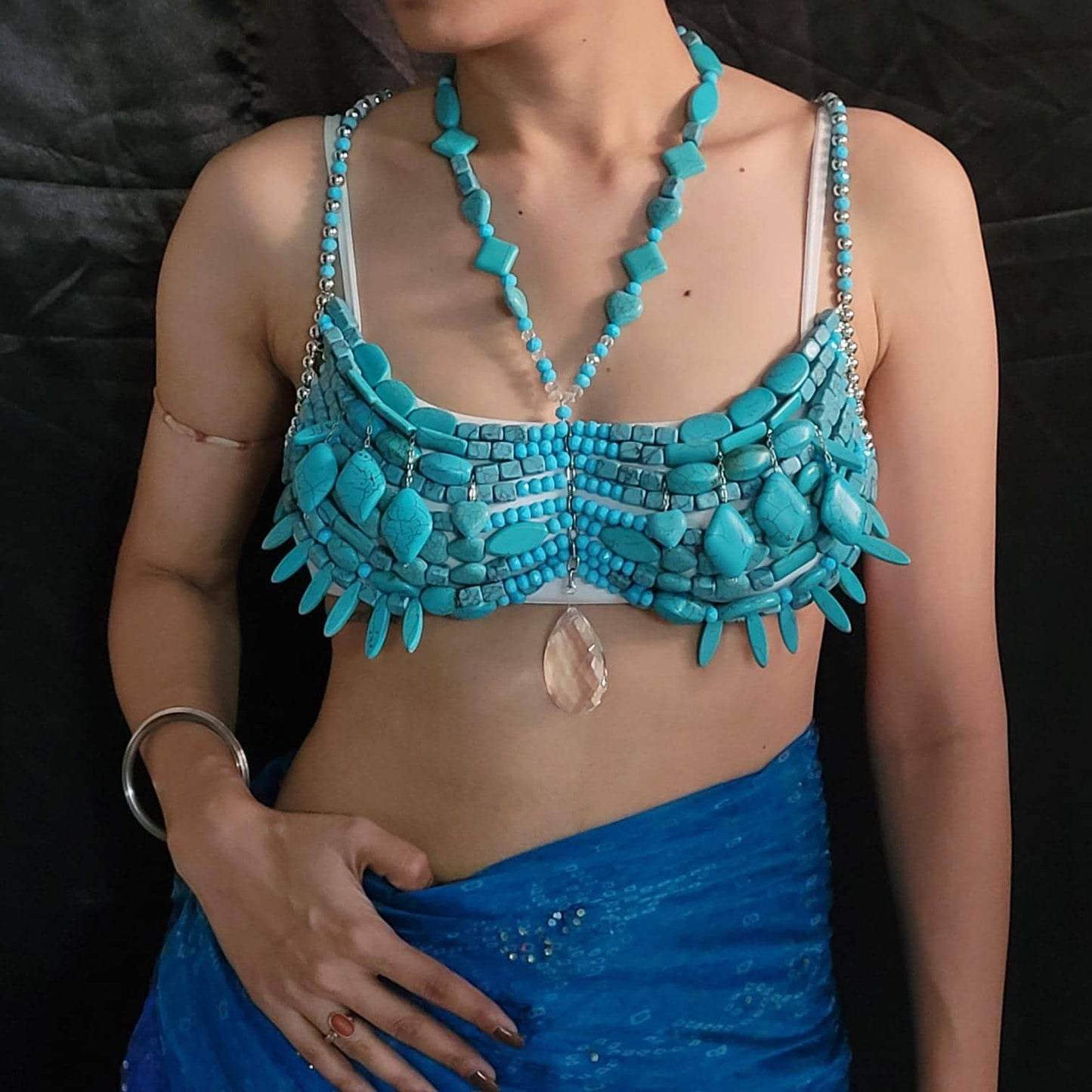 May include: A turquoise bra and necklace set. The bra is made of turquoise stones and has a unique design. The necklace features turquoise beads and pendants. A blue fabric is draped below the bra.