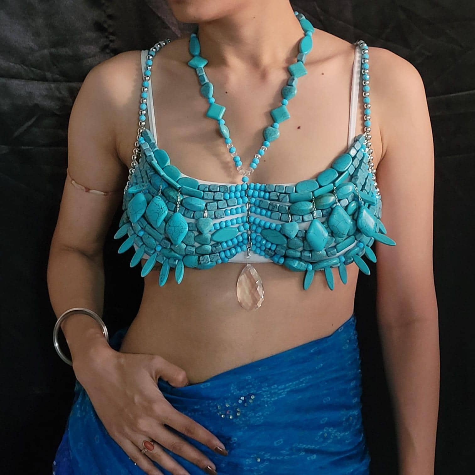 May include: A turquoise bra and necklace set. The bra is made of turquoise stones and has a unique design. The necklace features turquoise beads and pendants. A blue fabric is draped below the bra.