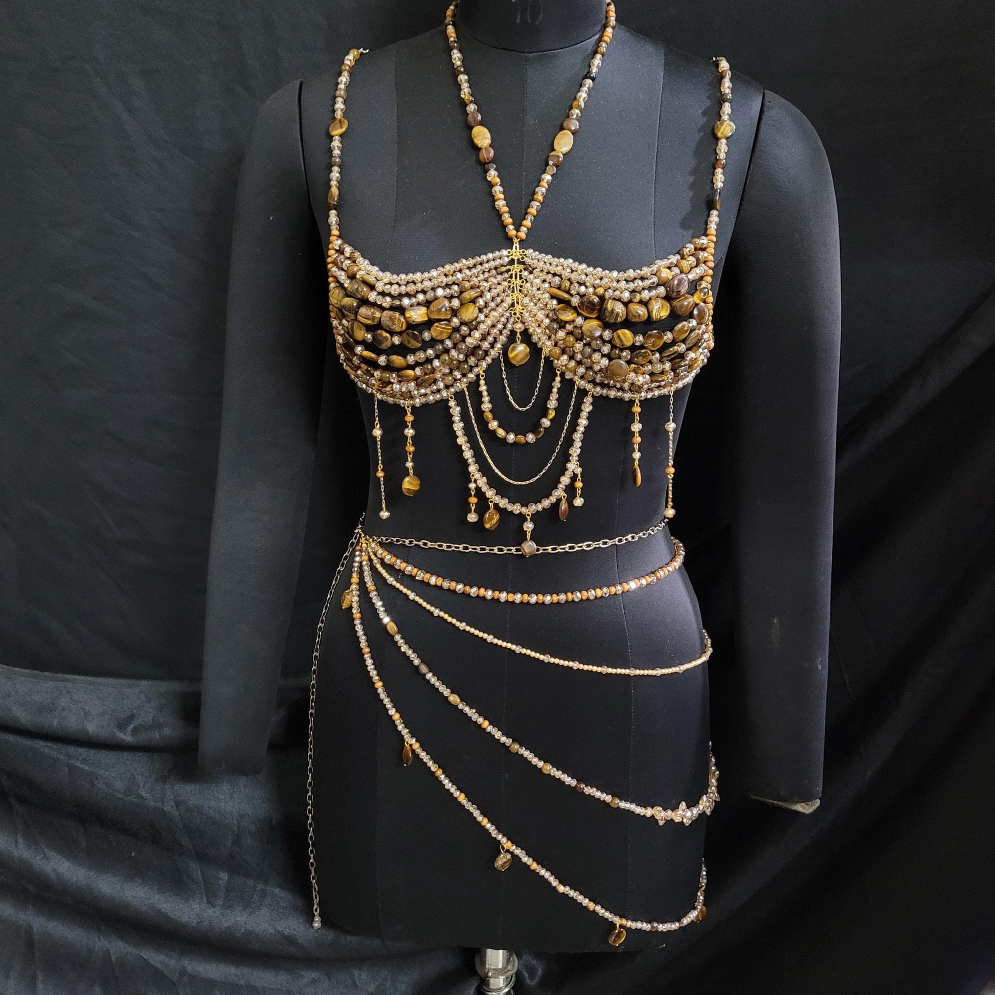 May include: A handcrafted, gold-toned body jewelry set on a black mannequin. The set includes a beaded bra, a necklace, and a multi-strand waist chain. The bra and necklace feature tiger's eye stones and small beads. The waist chain has multiple strands with dangling beads.